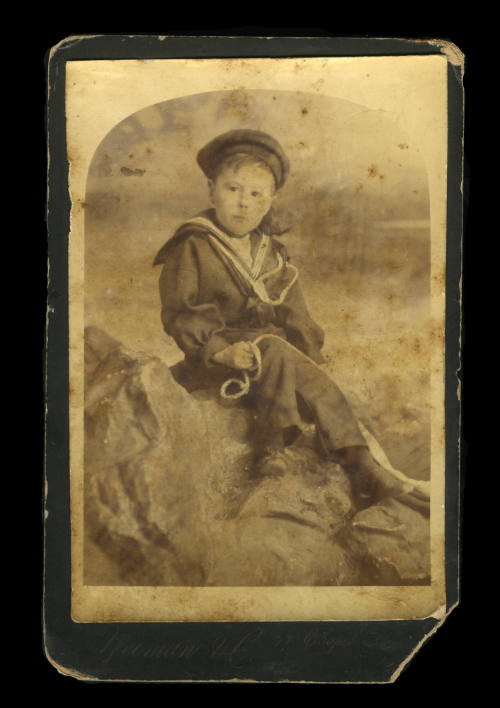 Young boy in a sailor outfit, holding onto a rope, and sitting on a rock