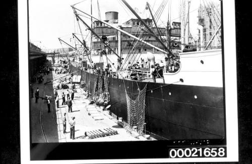 Wharf labourers using derricks to load bales of wool and crates into the holds of SS CANADIAN CRUISER 