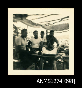 Photograph of four men, standing underneath a shade cover, on Packe Island