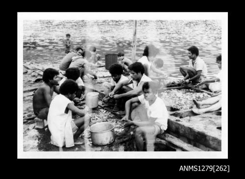 Photograph of men, women and children cleaning pearl shells, sitting around pearl shell cages, and with buckets and containers